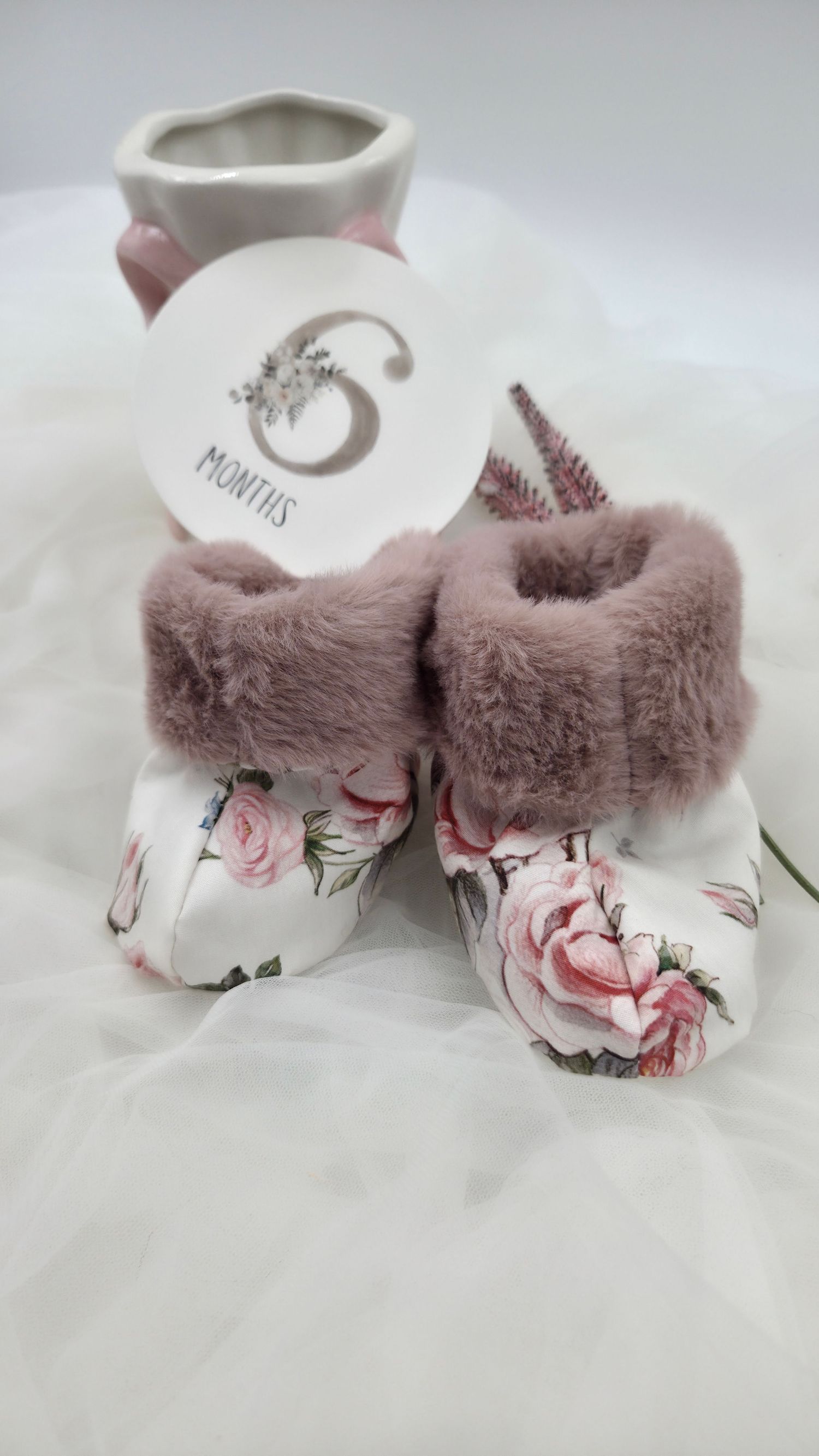 Floral baby booties with mauve faux fur trim displayed with a 6 months milestone card on white fabric.