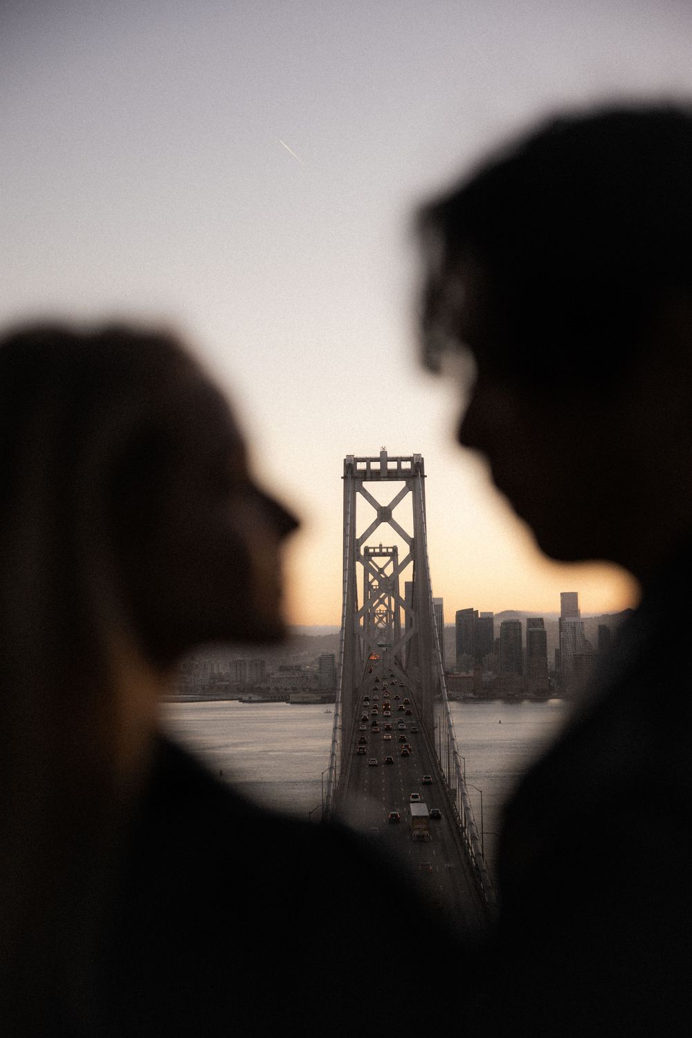 Silhouette of couple facing each other with San Francisco Bay Bridge and city skyline at sunset in background.