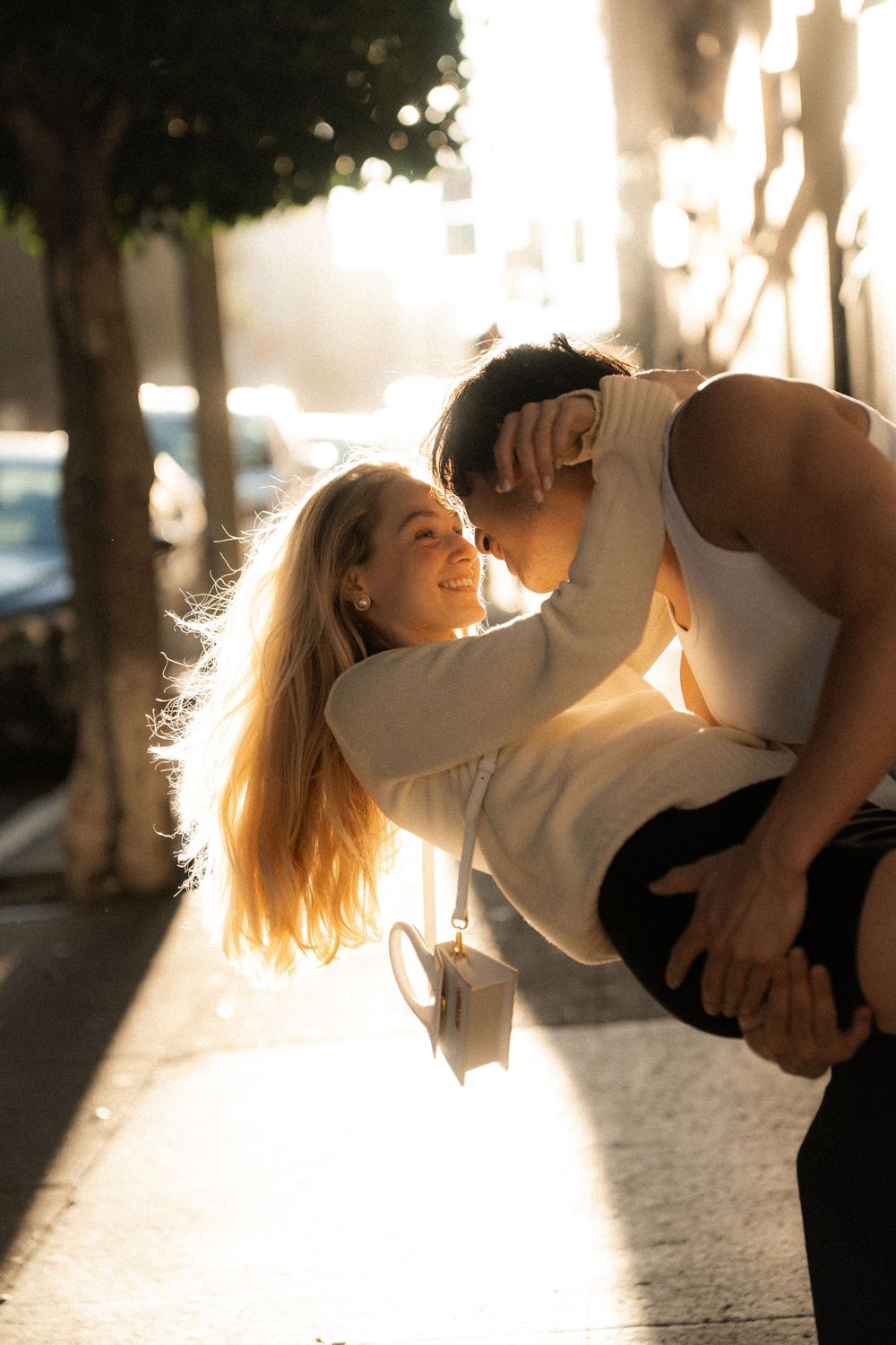A joyful couple shares a romantic moment on a sunlit sidewalk, with the man leaning in to kiss the smiling woman.