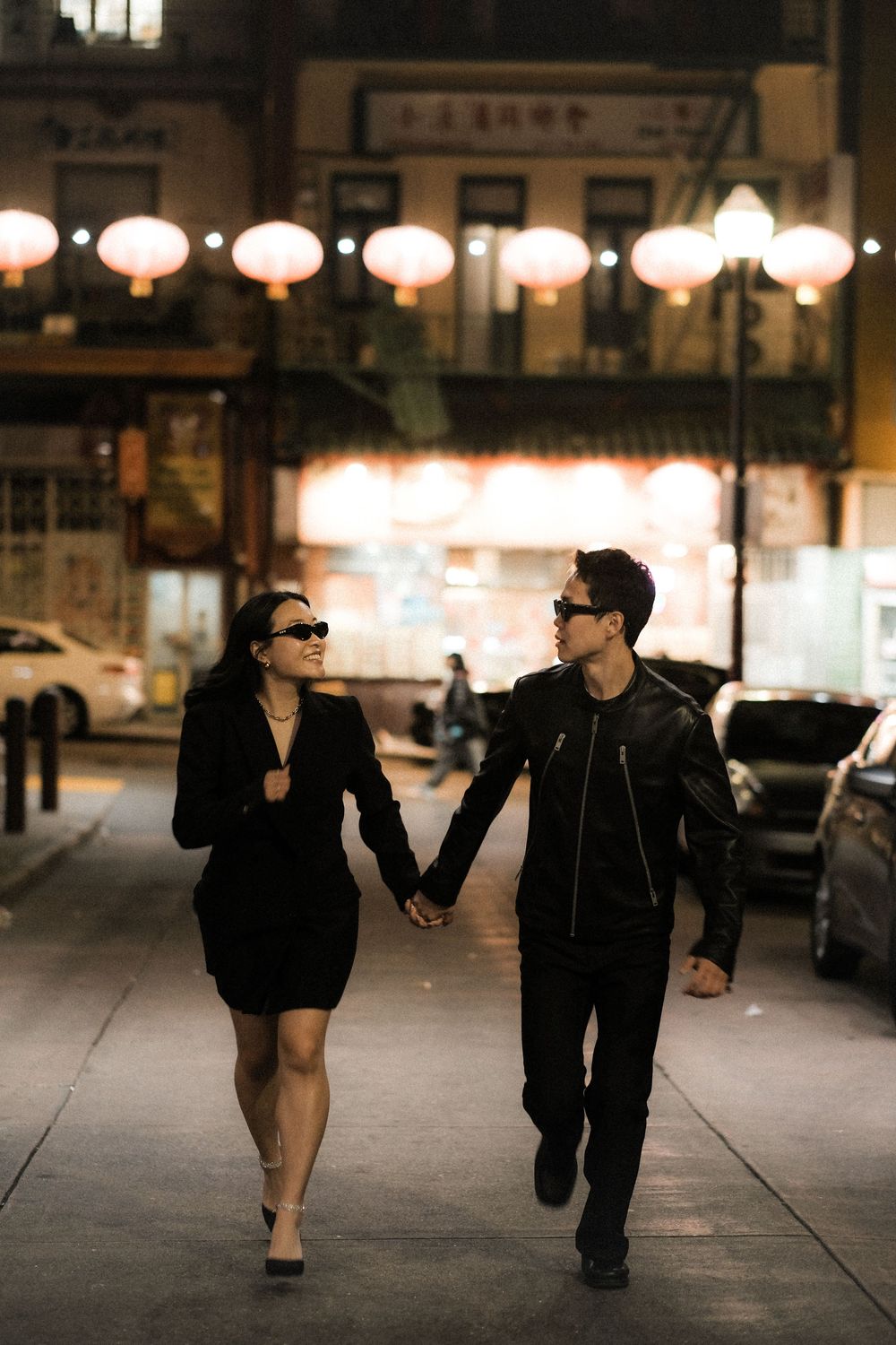 Two people walk together at night on a city street illuminated by glowing lanterns and storefront lights.