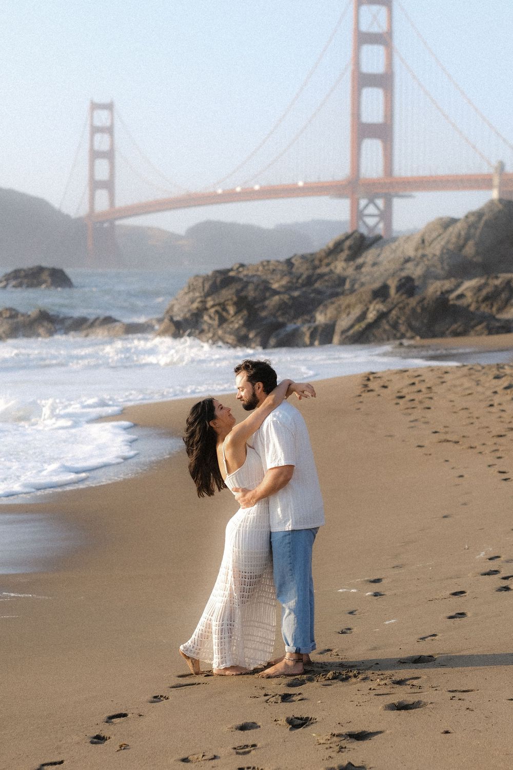 Romantic couple walking along Baker Beach with iconic Golden Gate Bridge in foggy San Francisco backdrop.
