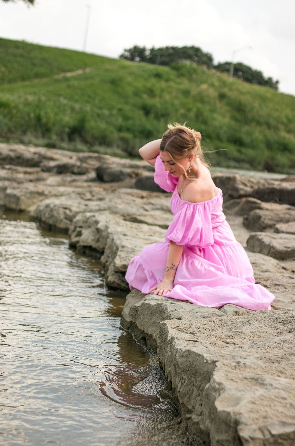 Woman in pink off-shoulder dress sitting on rocky shore, touching her hair near shallow water with green hills behind.