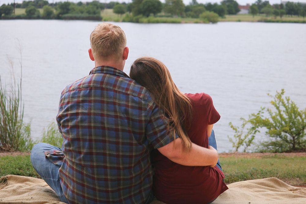 Couple sitting together by a peaceful lake, embracing while enjoying the scenic waterfront view on a sunny day.