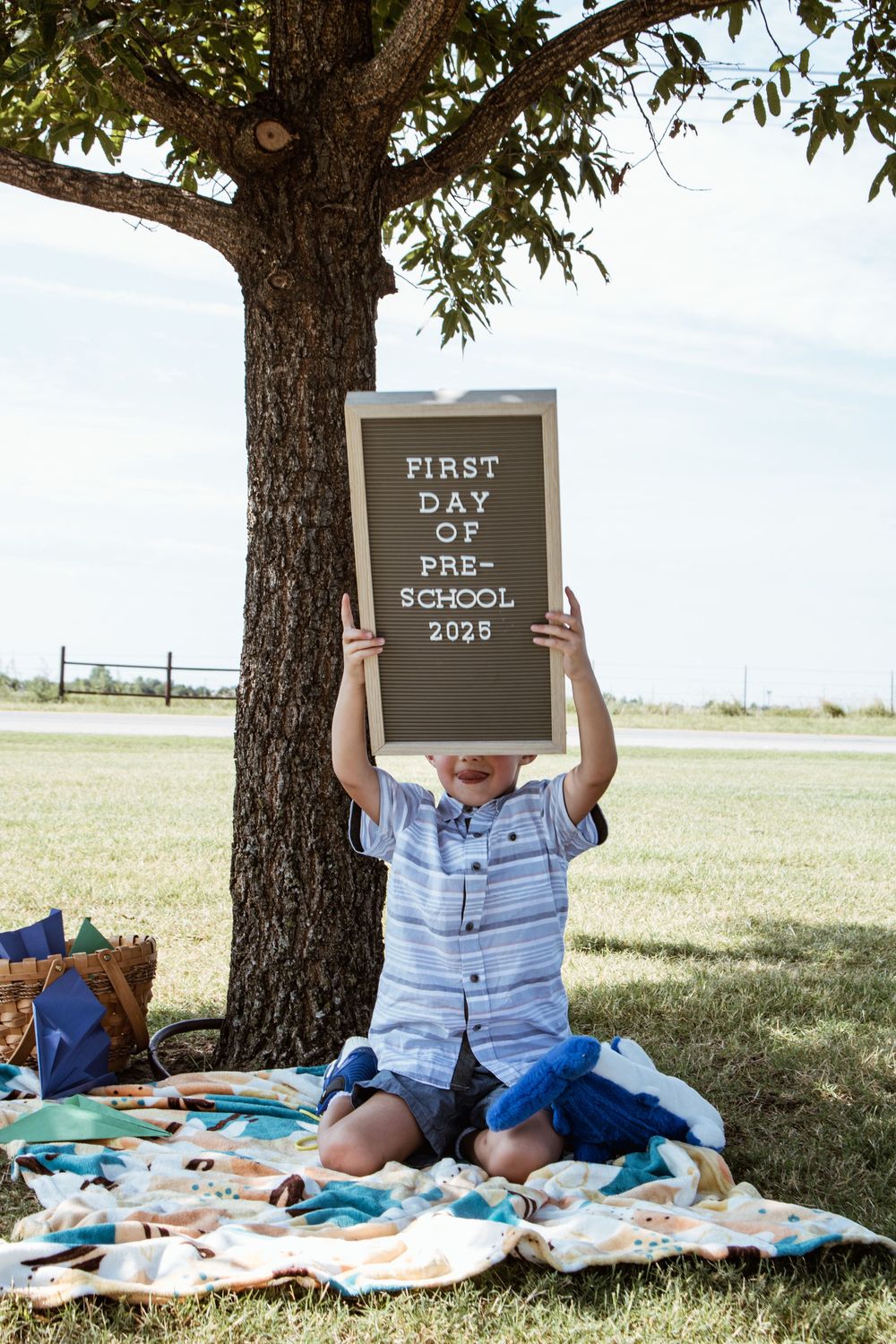 Child with chalkboard signs announcing first day of preschool 2023 under tree.
