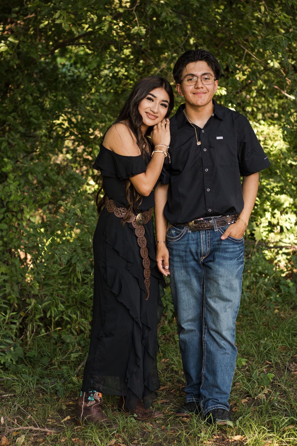Couple in black and denim poses against lush greenery in romantic outdoor photoshoot.