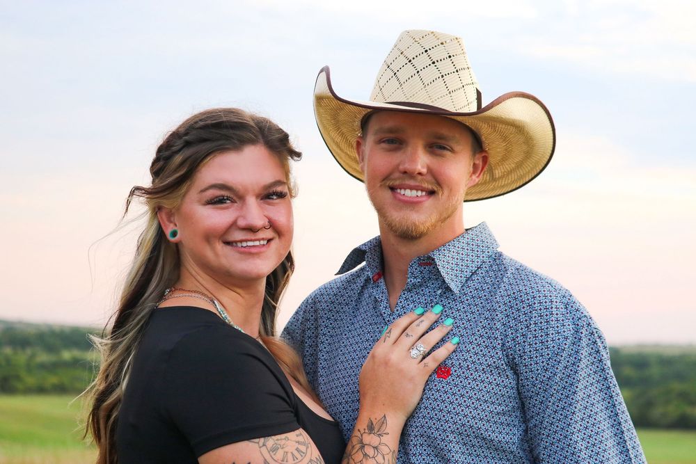 A smiling couple poses outdoors, woman with tattoos in black top, man in cowboy hat and blue plaid shirt.