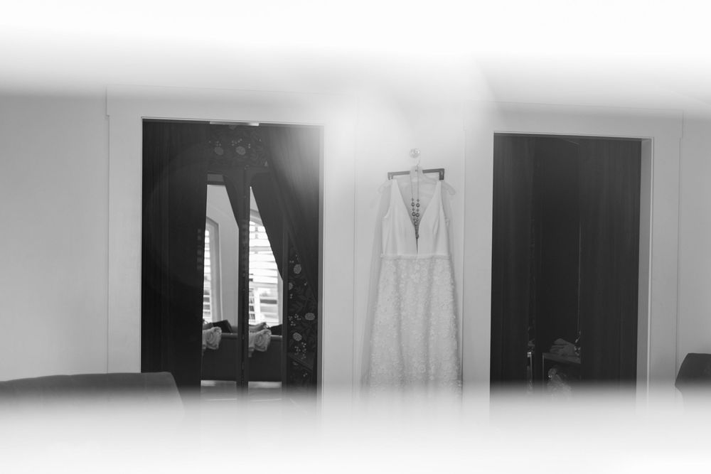 Black and white photo of an elegant wedding dress hanging in a bright room with dark wooden doors.
