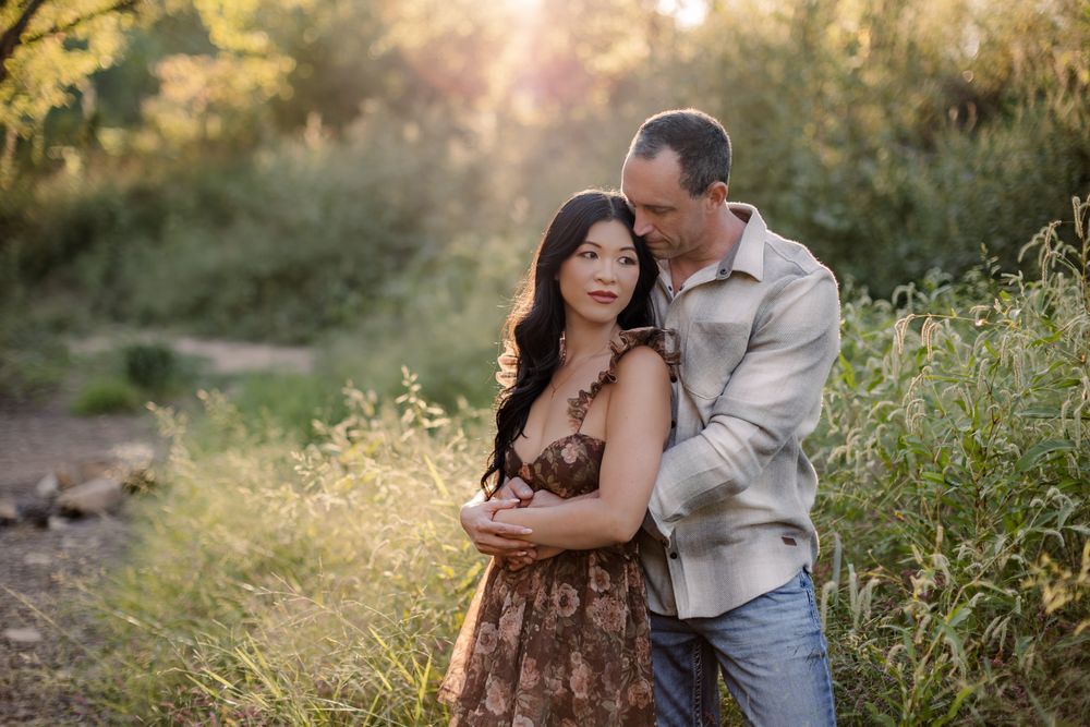 Couple embracing in golden hour sunlight among tall grass and wildflowers in a romantic outdoor engagement photo.