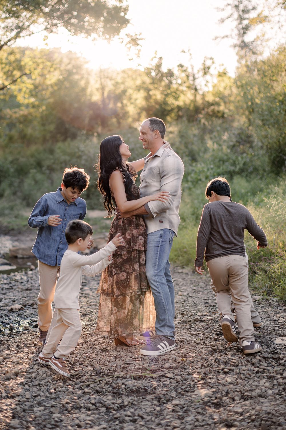 Family of five posing outdoors at golden hour, parents embracing while three boys play around them on rocky ground.