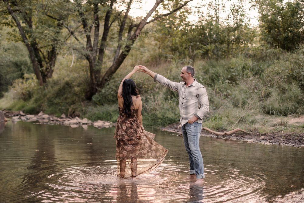 Couple dancing romantically in a shallow river surrounded by lush green trees during golden hour.