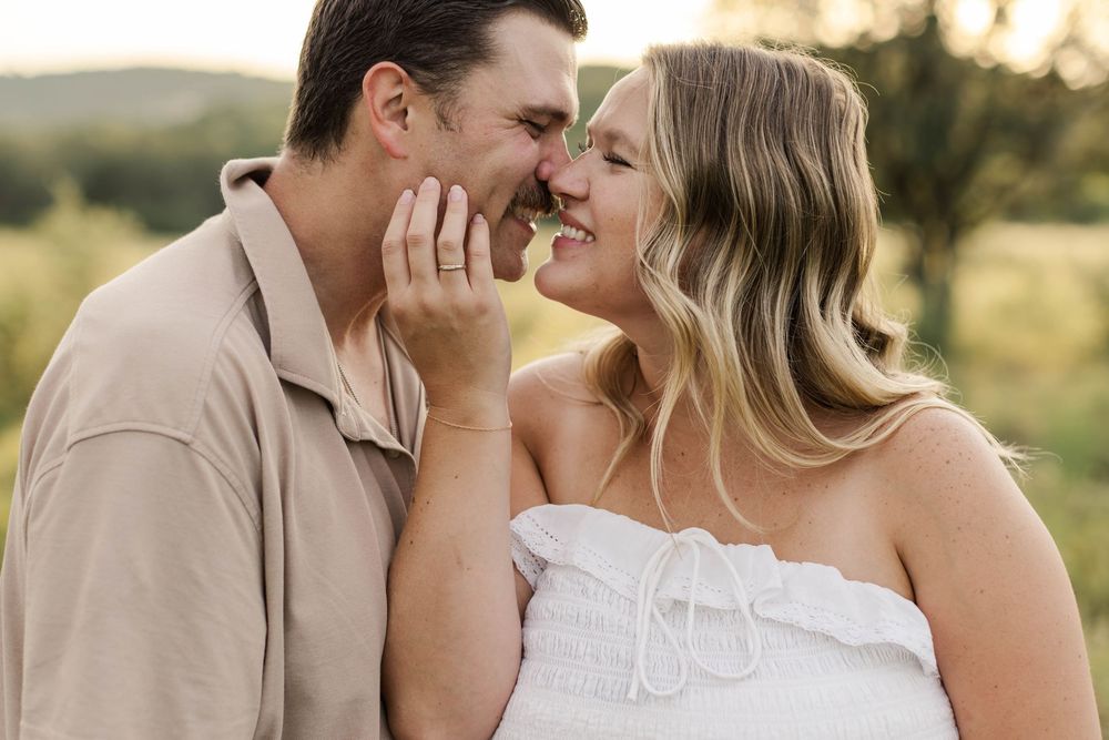 A couple shares a romantic moment outdoors, smiling and touching foreheads in a lush green setting.