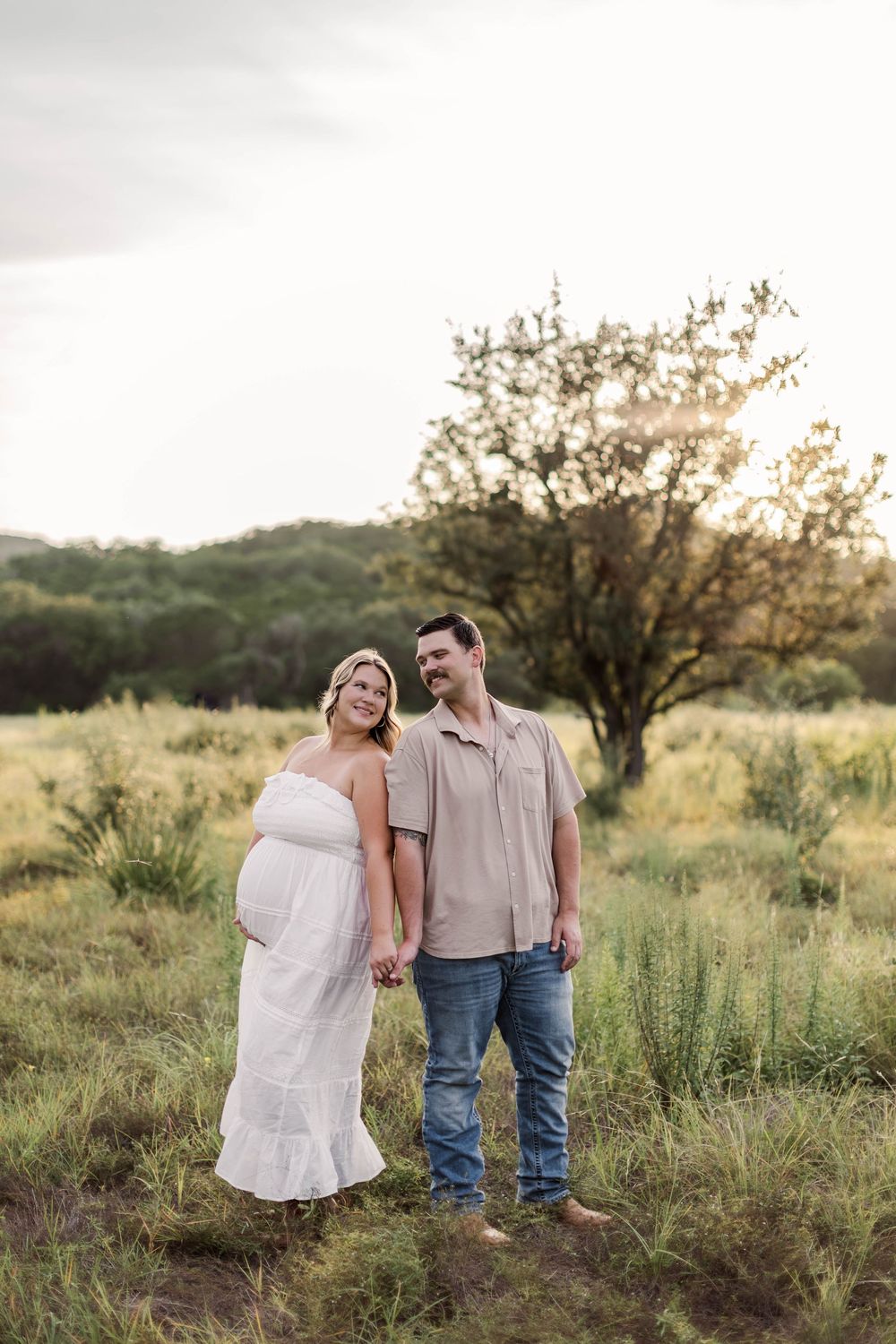 Couple holding hands in a green field at golden hour, woman in white maternity dress, man in beige shirt and jeans.