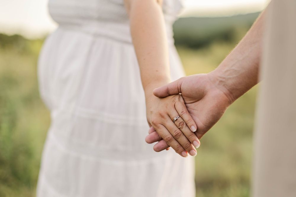 Couple holding hands outdoors, woman wearing engagement ring and white dress, romantic field background.