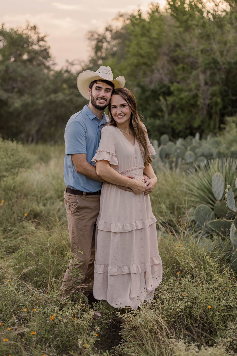 A couple poses in a Texas field at sunset, man in cowboy hat and blue shirt, woman in blush ruffle dress.