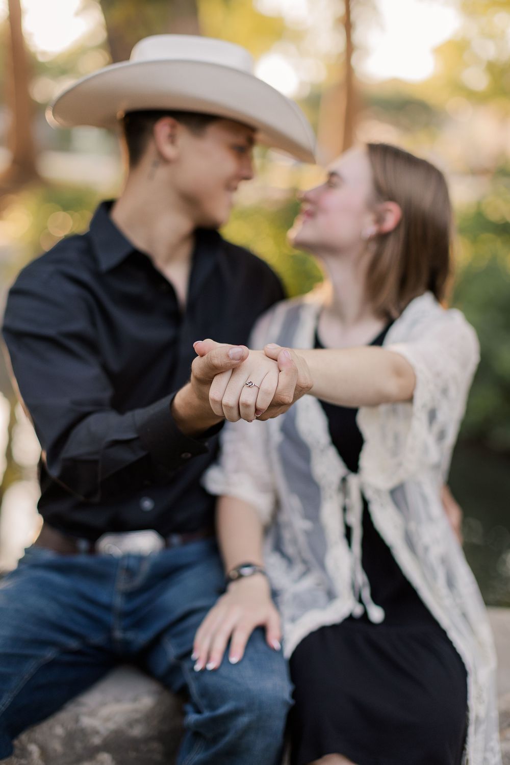 Couple holding hands showing engagement ring, man in cowboy hat and black shirt, woman in lace cardigan, outdoors.