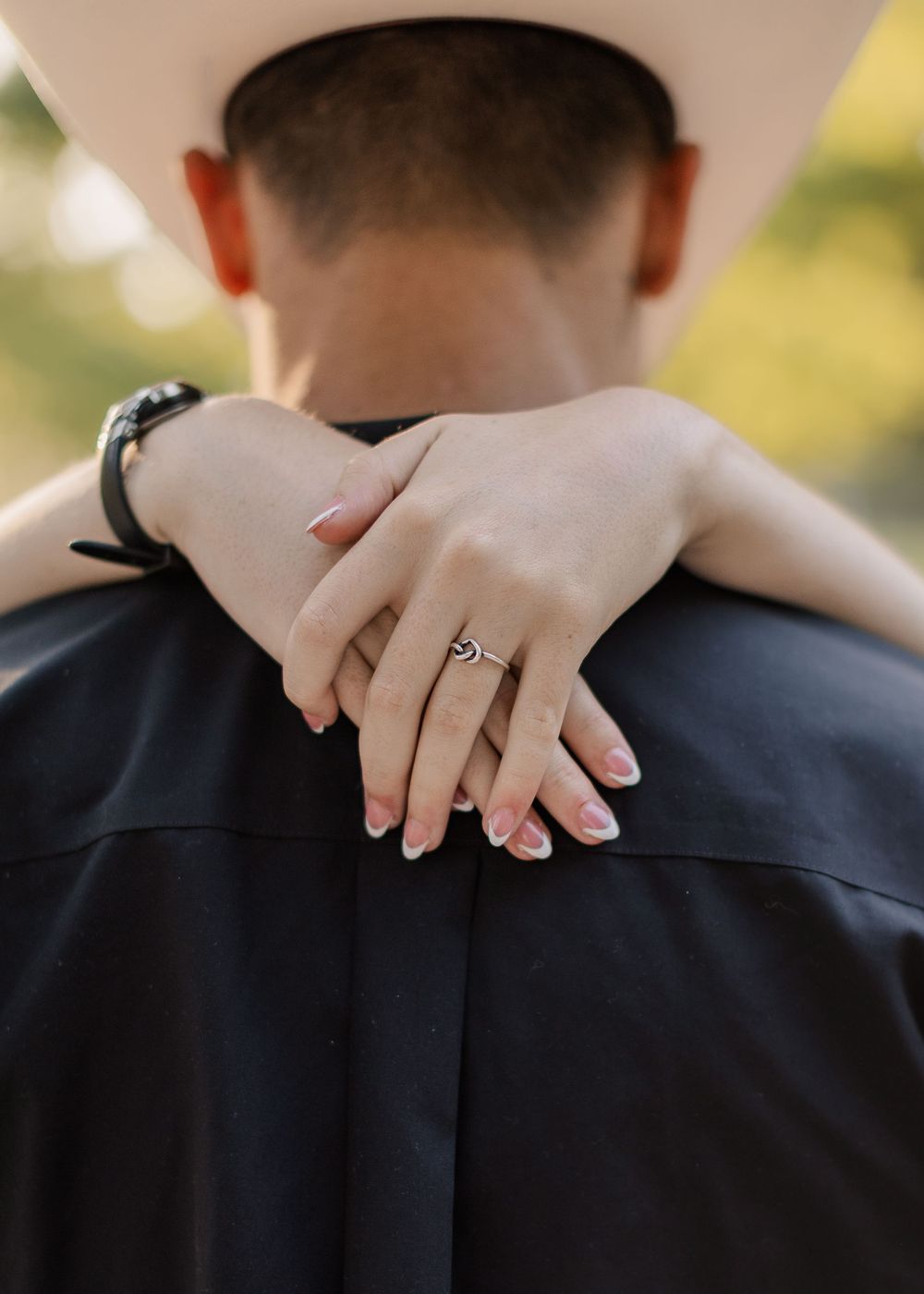 Couple embracing, woman's hands clasped around man's neck showing engagement ring, wearing cowboy hat, outdoor setting.