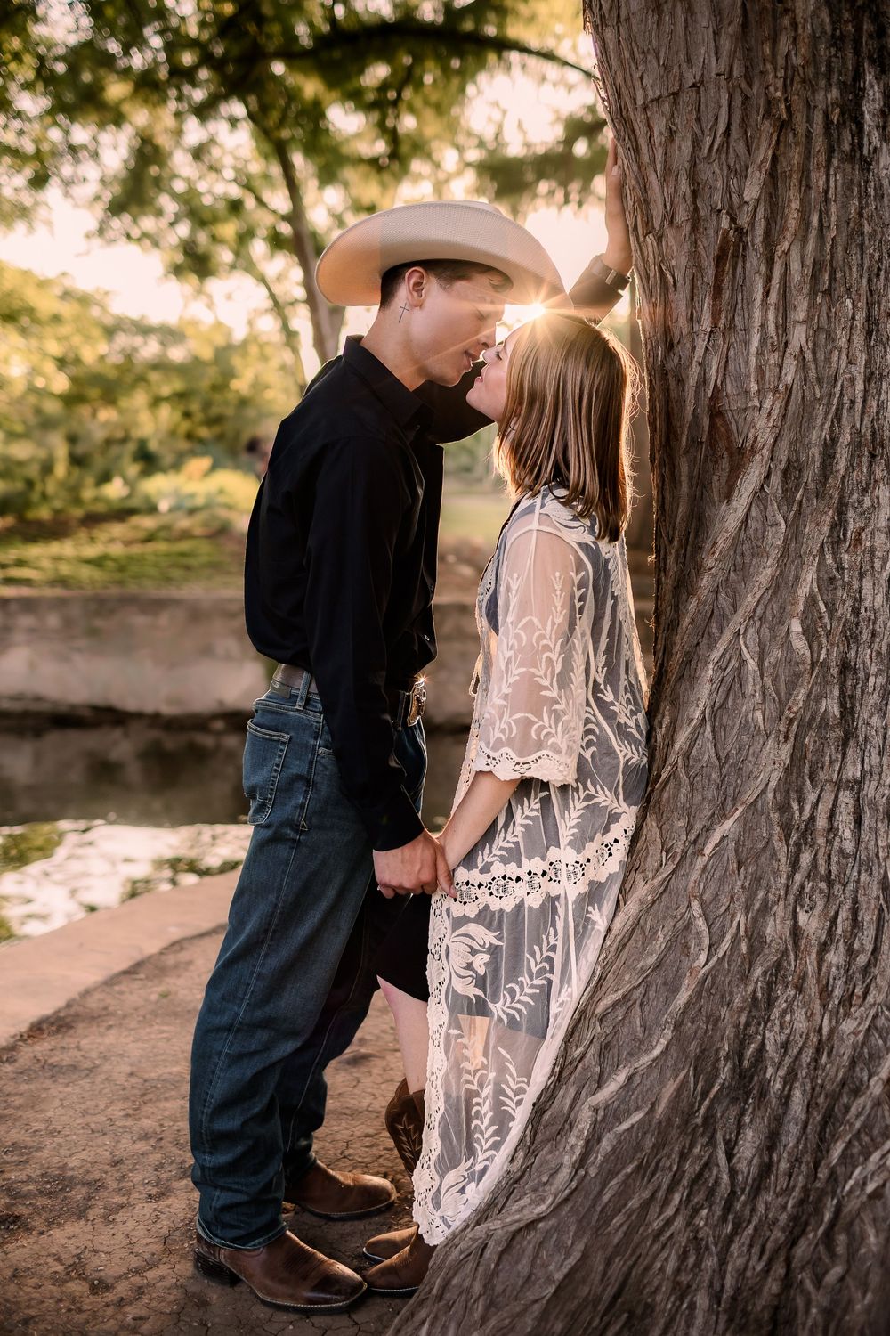 Couple sharing a romantic moment near a tree at golden hour, man in cowboy hat and woman in lace dress holding hands.