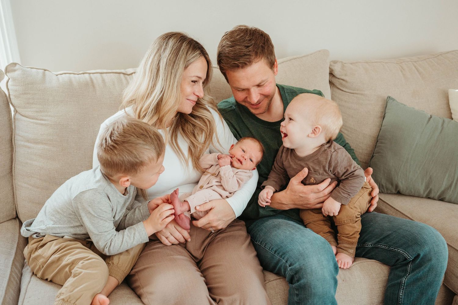 Happy family of five with two young boys and a newborn baby girl cuddles together on a beige sofa.