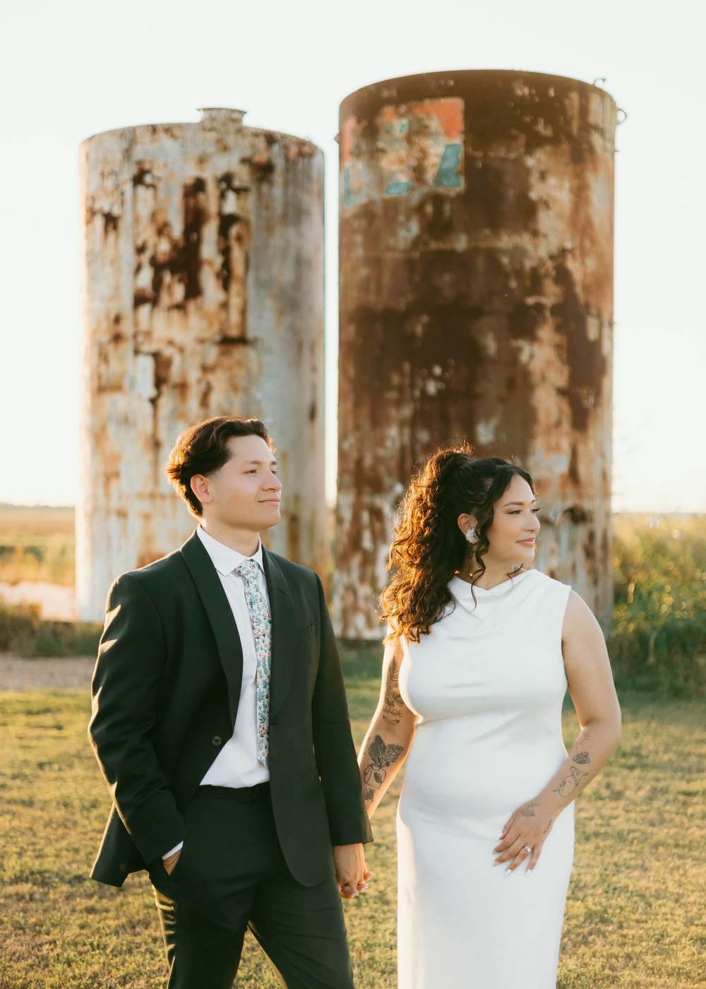 A couple in formal attire playfully poses in front of rustic metal silos during a sunset photoshoot.