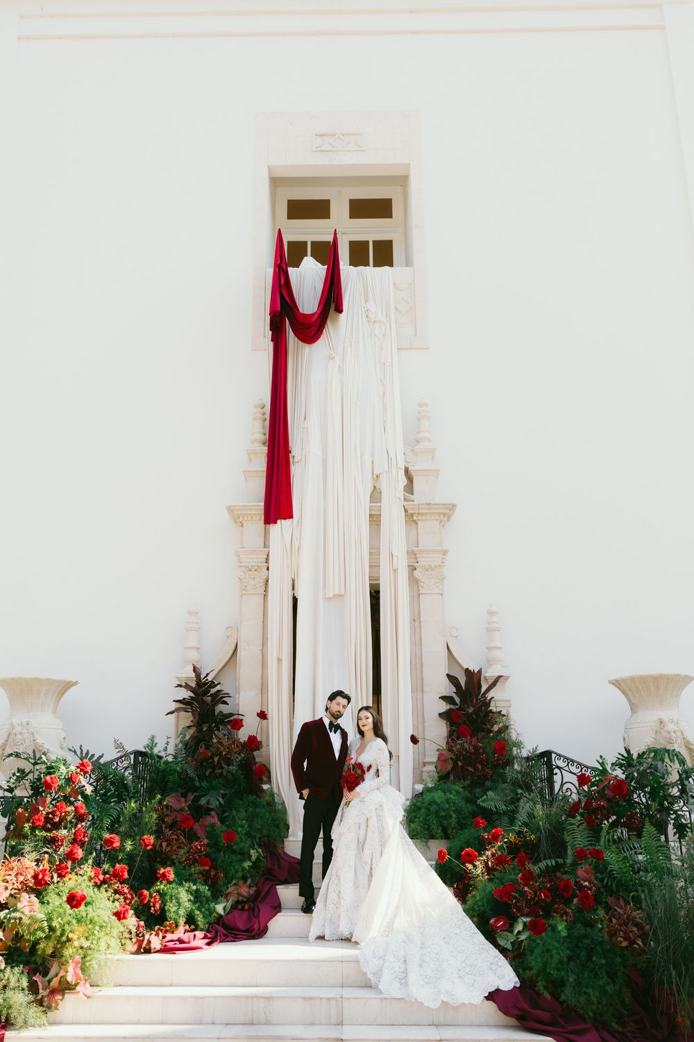 A romantic wedding scene unfolds on a white columned patio surrounded by vibrant red and orange flowers.