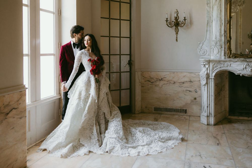 A bride and groom embrace in a romantic vintage room setting with ornate walls and large windows.