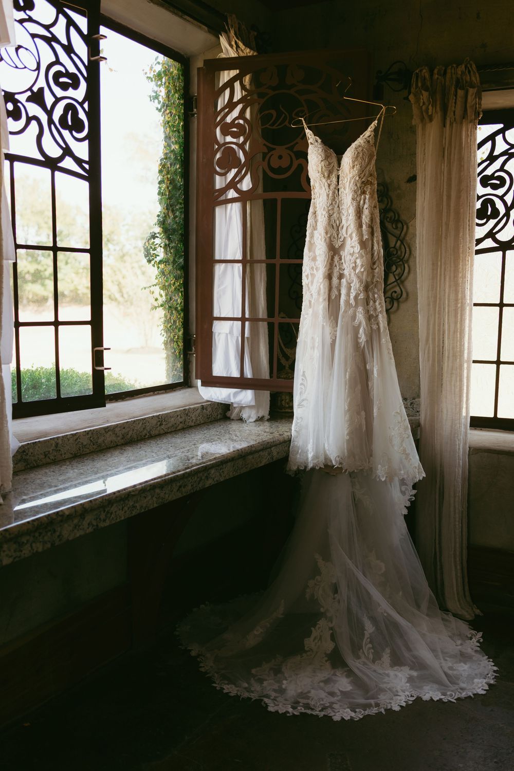 Elegant white wedding dress hanging by a rustic window in a vintage-style room with natural light streaming in.