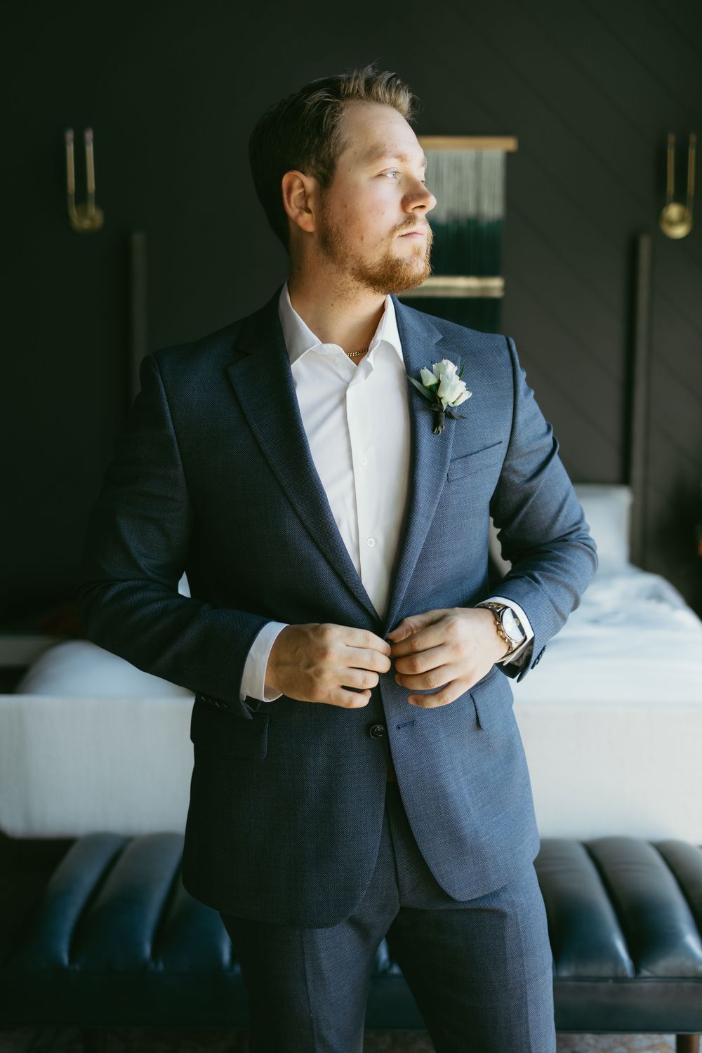 A groom in a navy suit adjusts his jacket while getting ready in a room with moody lighting and dark furniture.