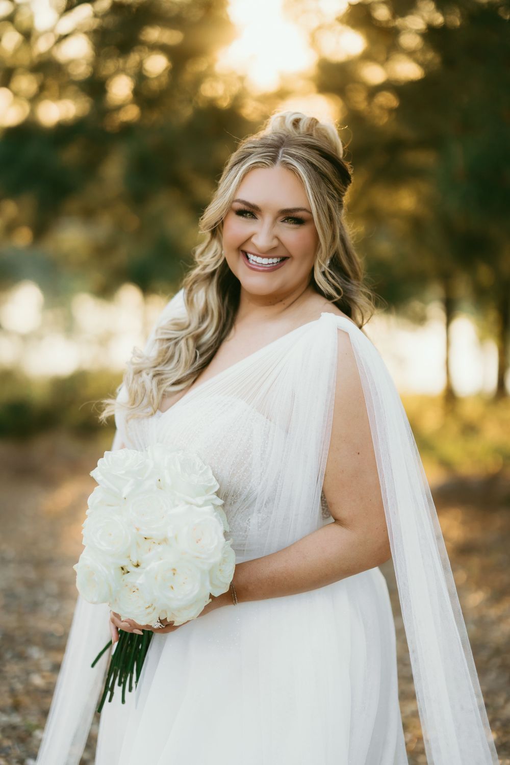 Bride in flowing white wedding dress holds white bouquet during golden hour sunset photography session.