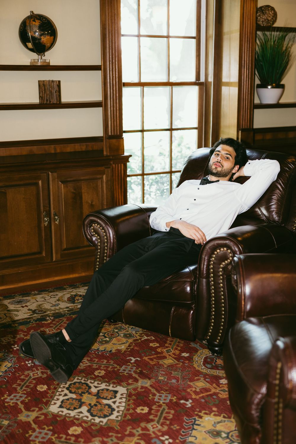 Person lounging in a brown leather armchair in a wood-paneled study with oriental rug and traditional decor.