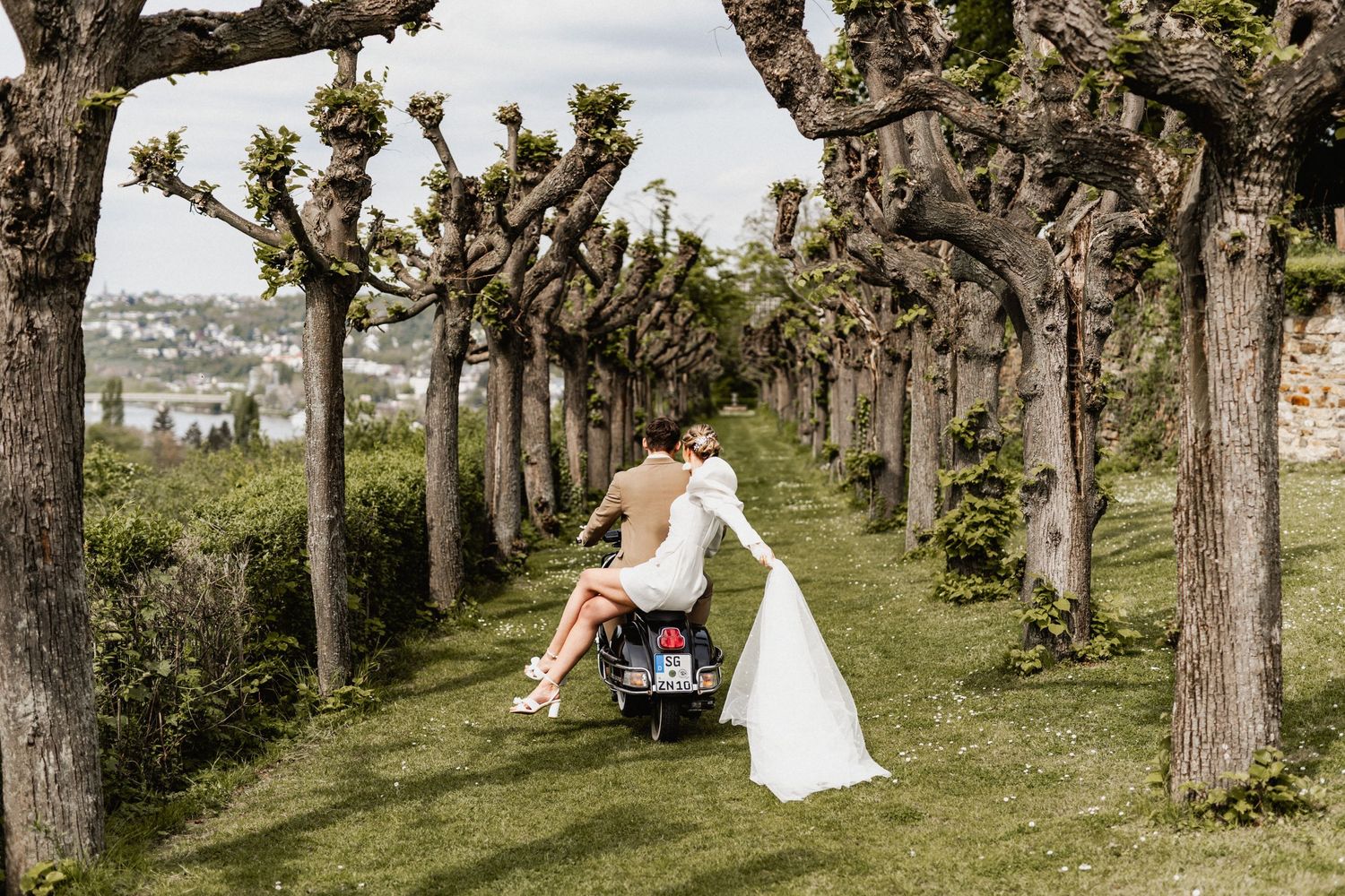 A couple poses with their Vespa scooter along a scenic tree-lined pathway.