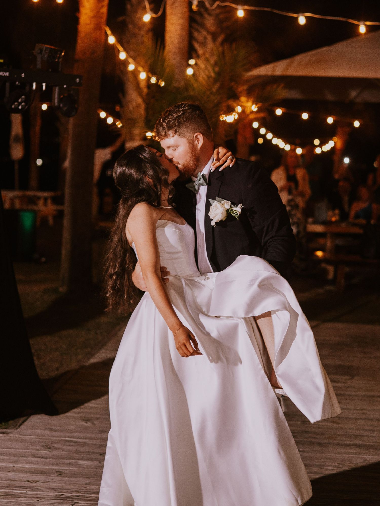 Couple in formal wedding attire dancing under string lights at night.