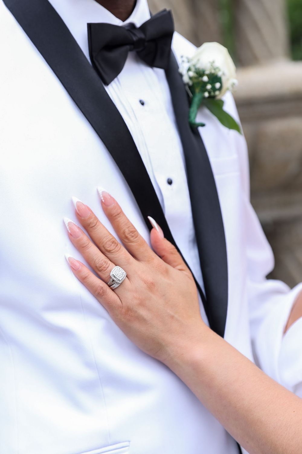 Close-up of a diamond engagement ring with white tuxedo lapel and boutonniere in background.