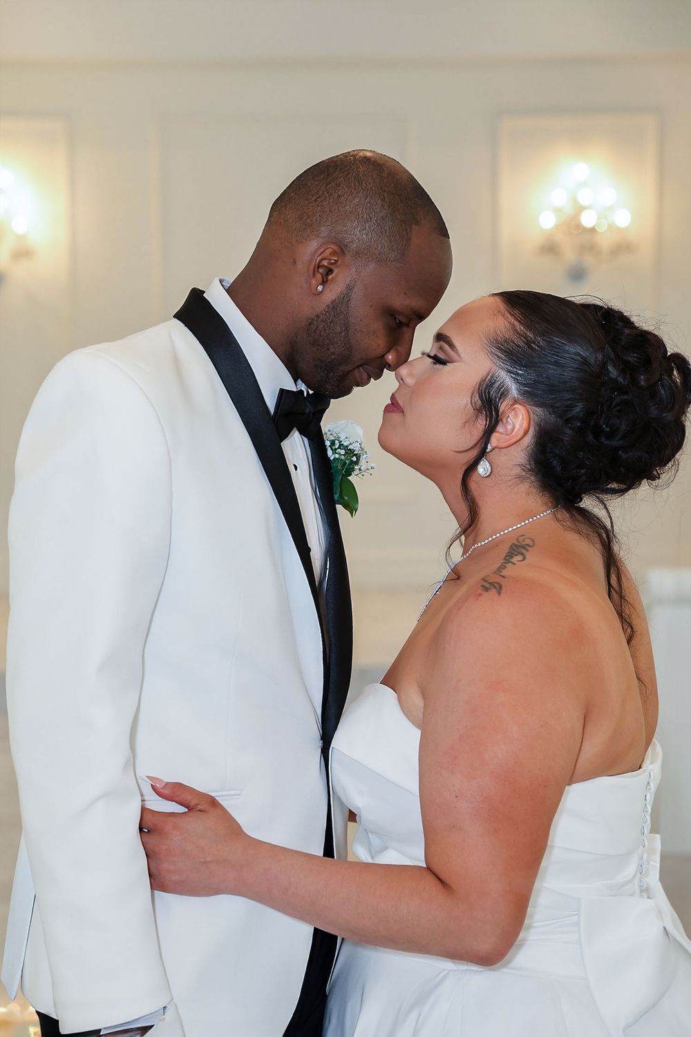 Intimate wedding moment in white formal attire against an elegant light-filled backdrop.