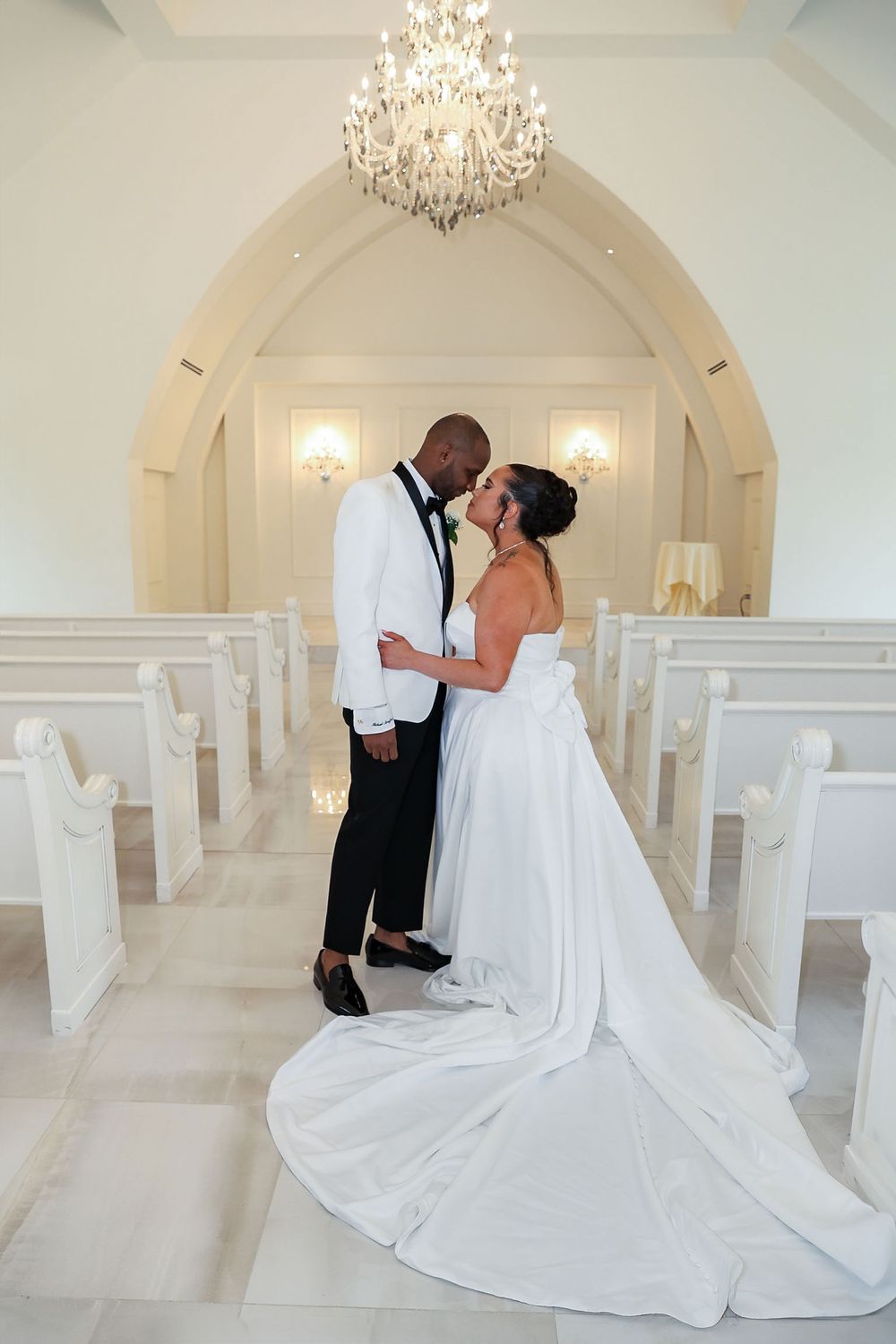 Romantic wedding photo in all-white chapel with crystal chandelier and elegant decorative arch.