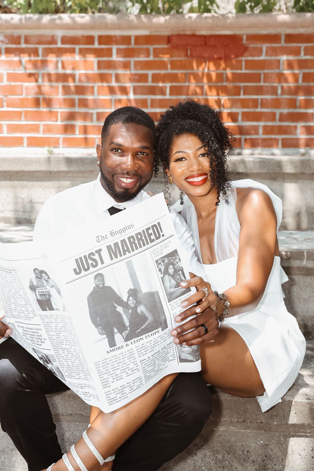 A couple in white outfits pose with a 'Just Married' newspaper against a brick wall.