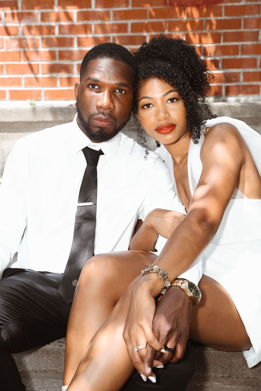 A couple in matching white and black attire pose intimately against a brick wall.