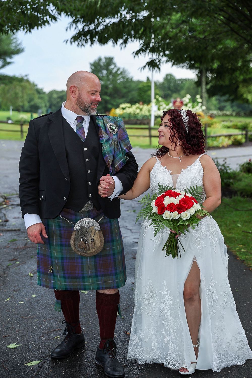 Wedding couple outdoors with groom in traditional Scottish kilt and bride holding red and white bouquet.