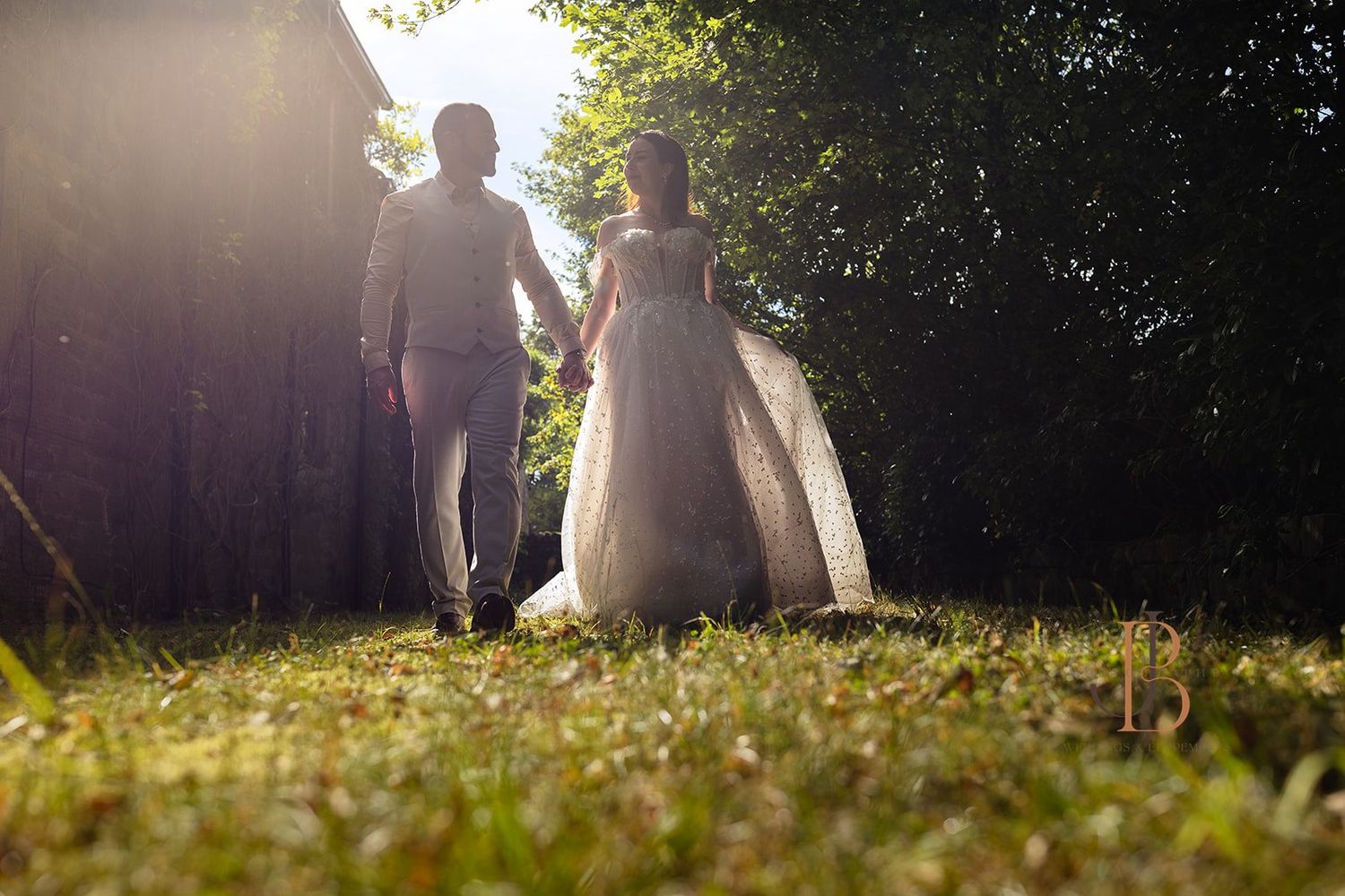 A bride and groom hold hands walking toward sunlit trees in a lush green garden on their wedding day. Yorkshire wedding photographer