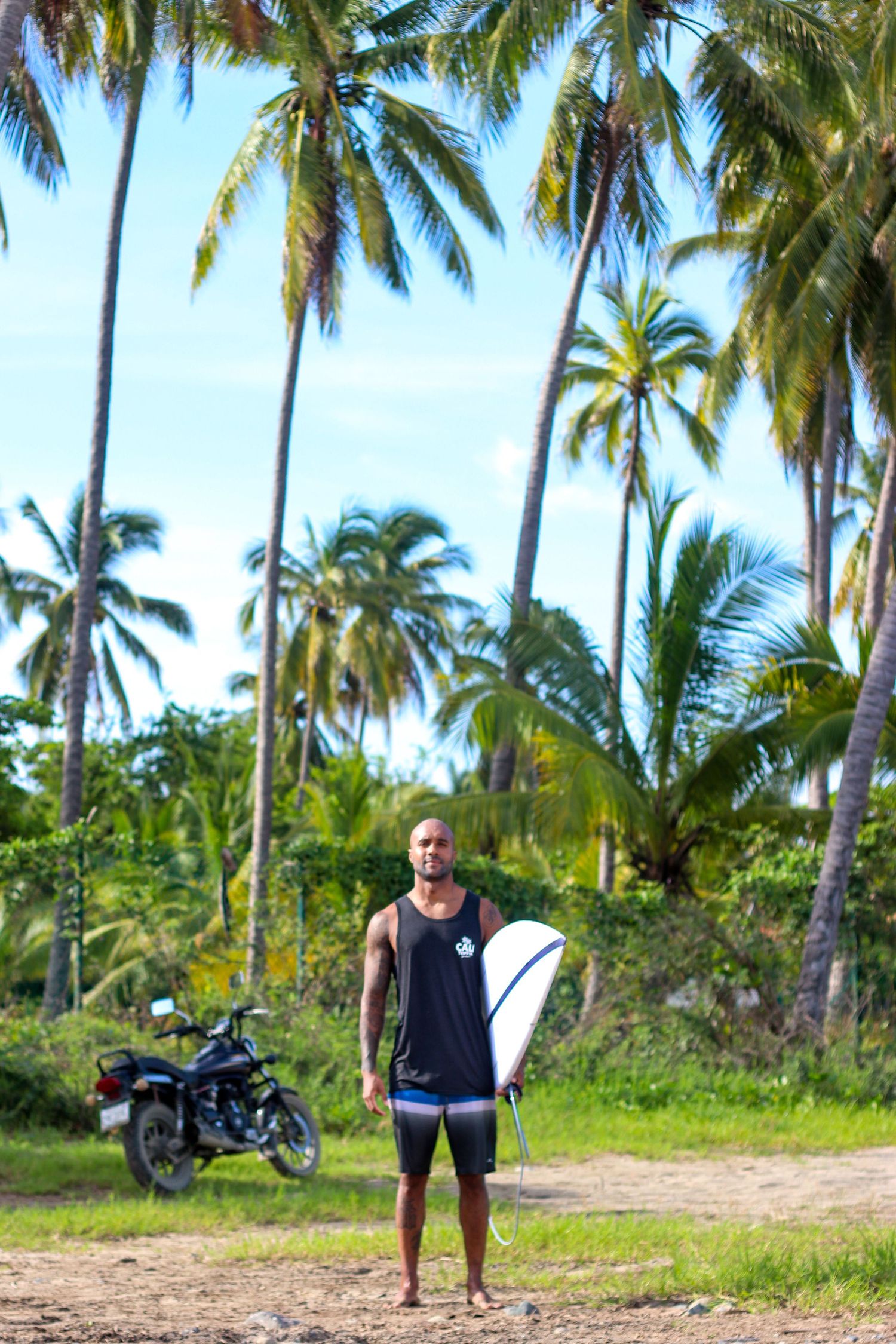 Tropical surf travel photography of a male surfer holding a surfboard, standing in front of lush palm trees and a motorcycle.