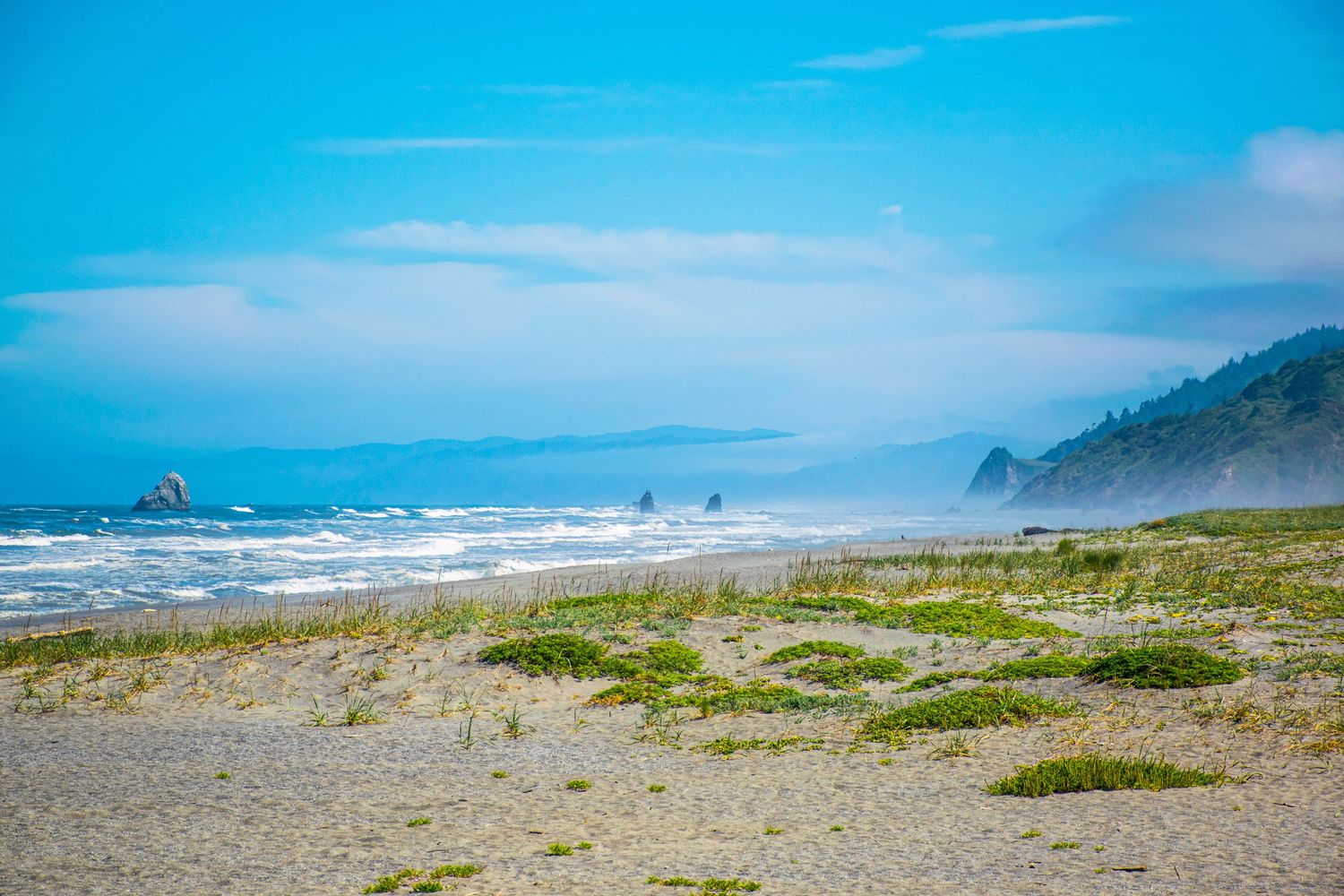 Foggy coastline with waves crashing on a sandy beach beneath blue skies.