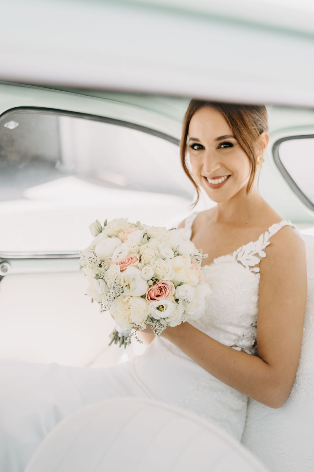 A bride sits in vintage car holding a soft pink and white wedding bouquet.