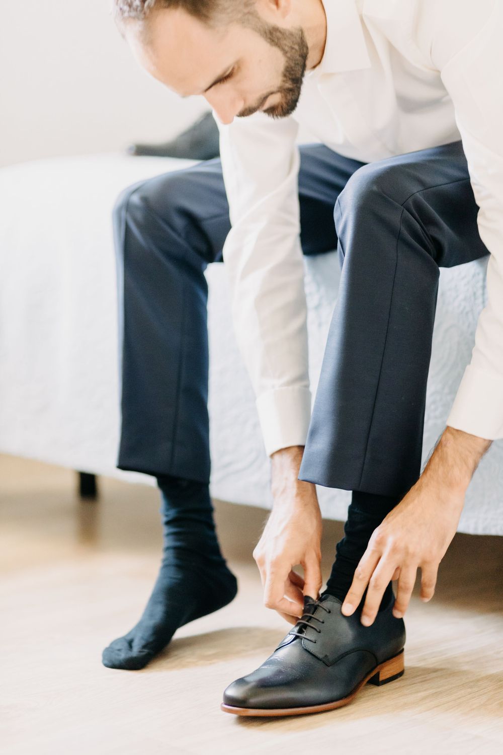 Groom in formal attire ties his black leather dress shoes while getting ready.