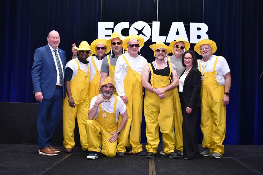 Group of people in yellow overalls and hats posing in front of an Ecolab banner at a corporate event.