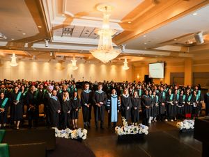 Large group of graduates in black and green robes pose together in an elegant banquet hall during graduation ceremony.