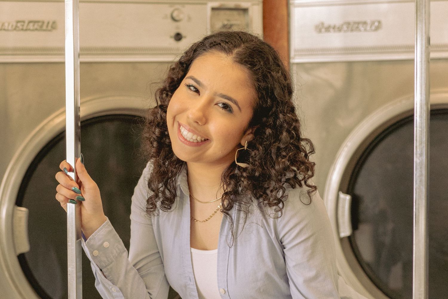 Young woman with curly hair smiling while leaning against washing machines in a laundromat.