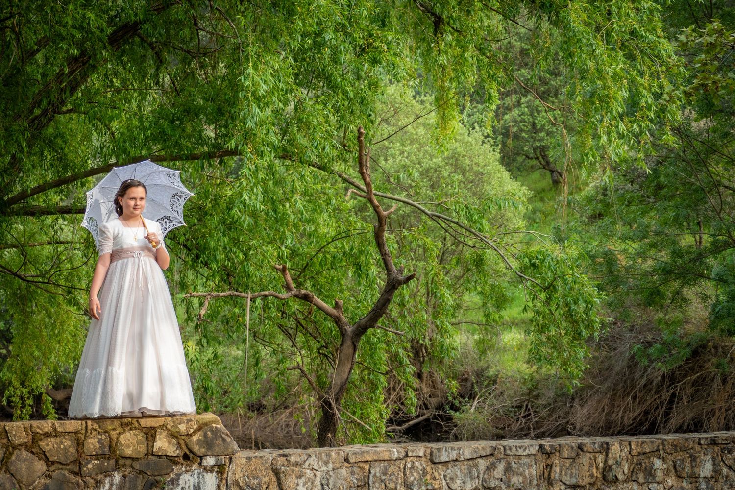 Bride in white gown holding lace parasol standing on stone wall surrounded by lush green trees.