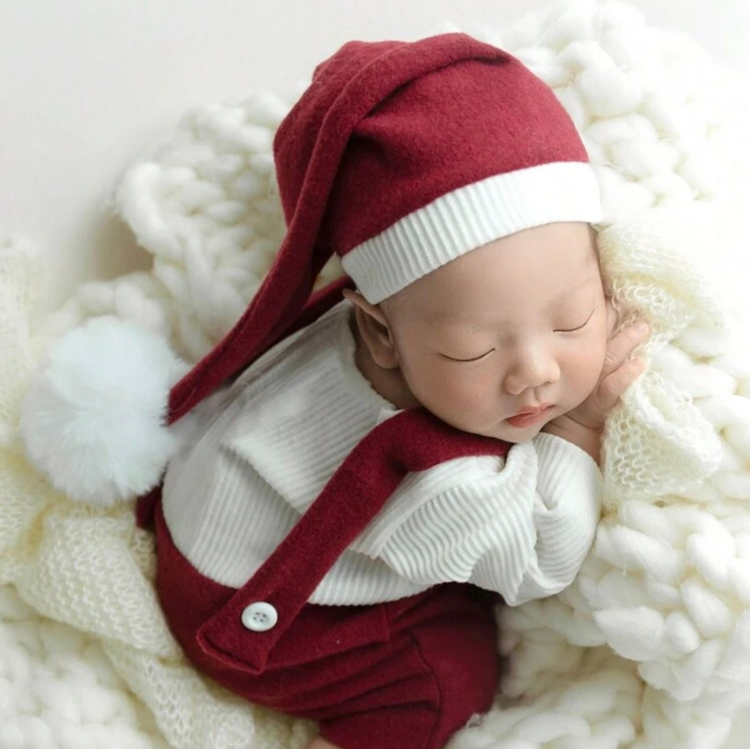 Baby sleeping on white fluffy surface wearing burgundy outfit with white collar.