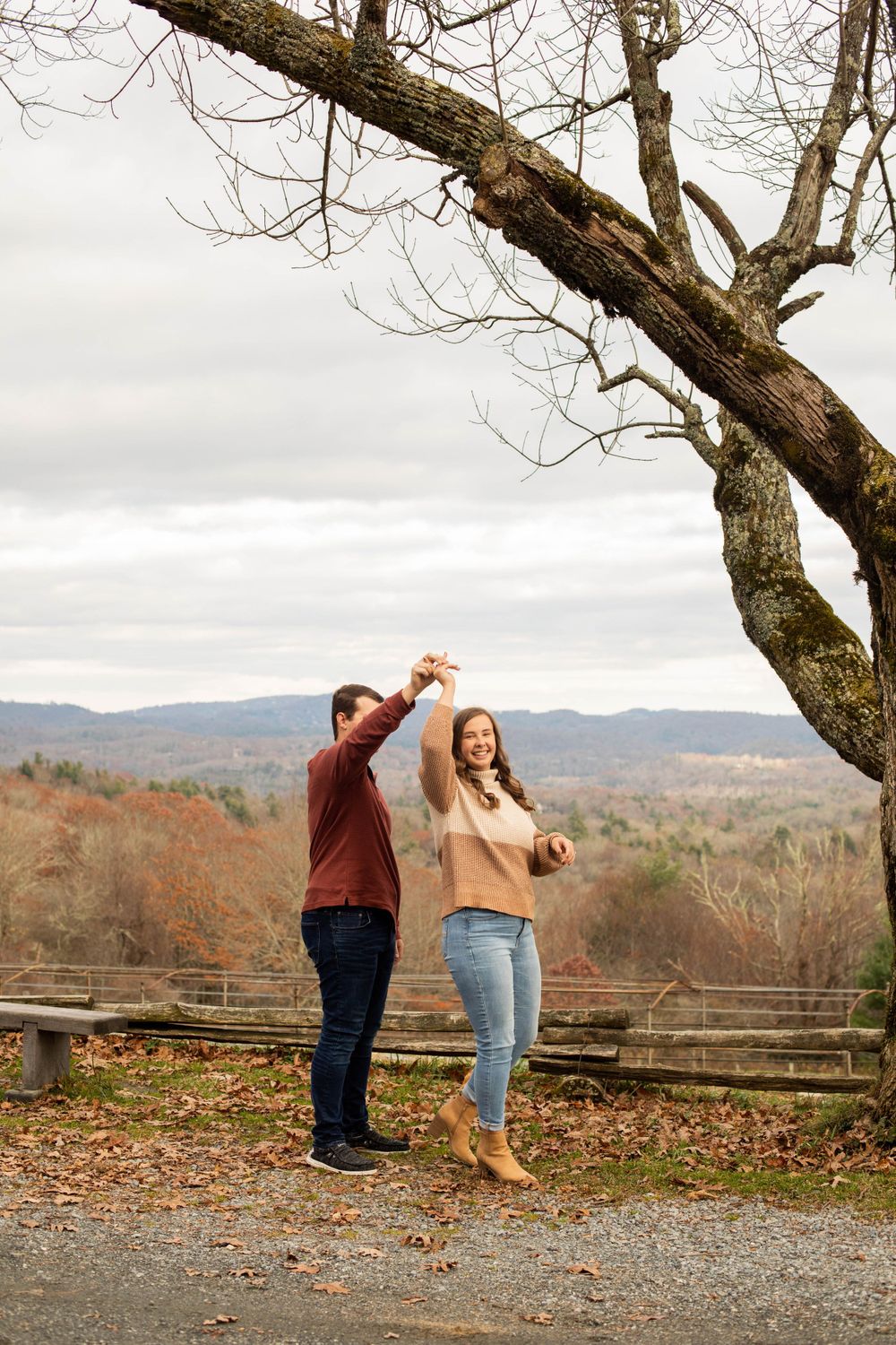 A couple dances playfully together on a scenic overlook with fall foliage in the background.