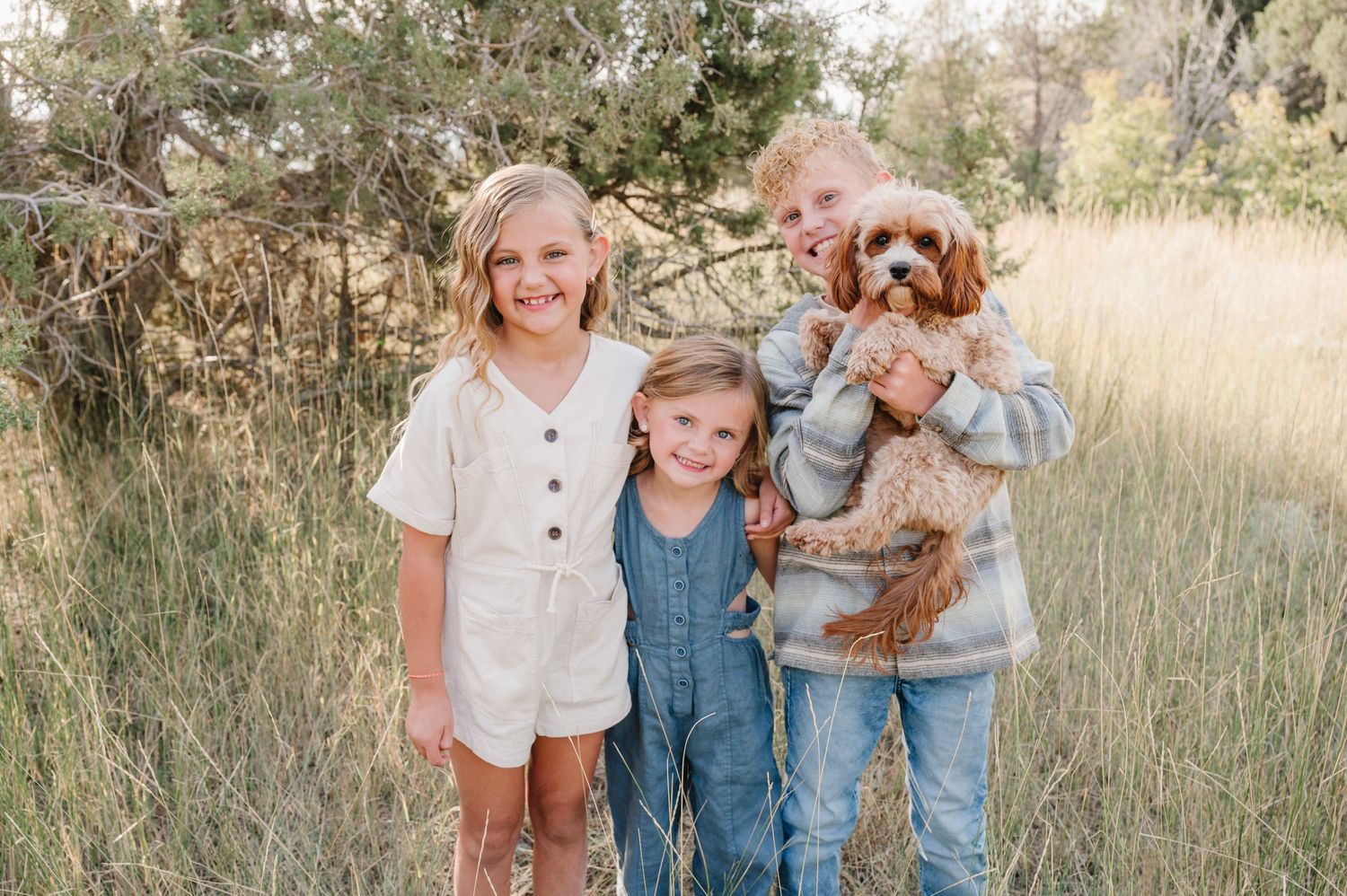 Three children in casual clothing pose together outdoors in a field with a small brown dog.