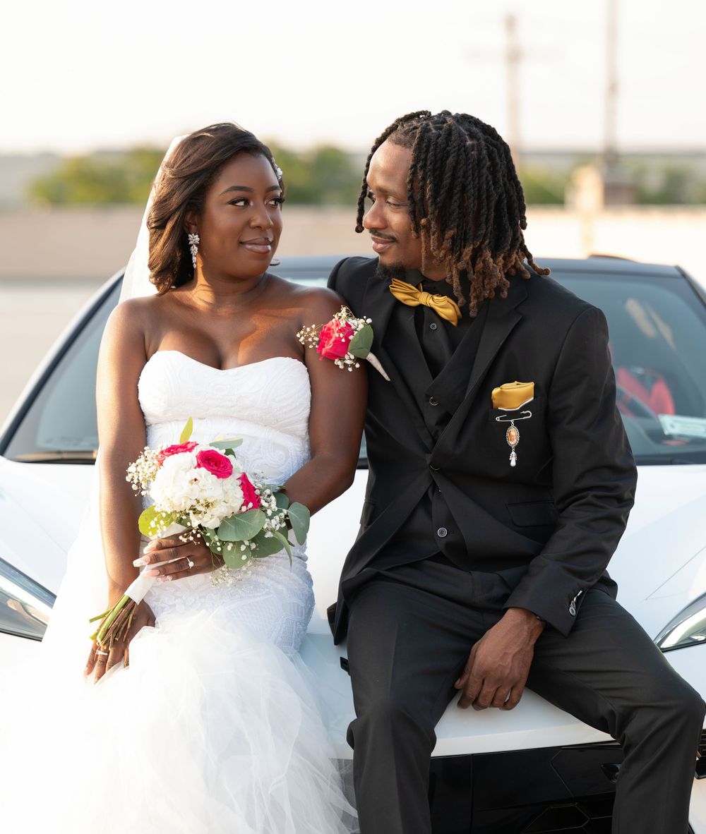 A newlywed couple poses against a white car, with the bride in white and groom in black formal attire.